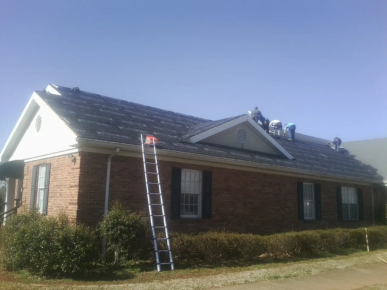 Crew tearing shingles off a brick ranch roof with a ladder set — Emergency Roof Repair work by our crew in South Bloomingville