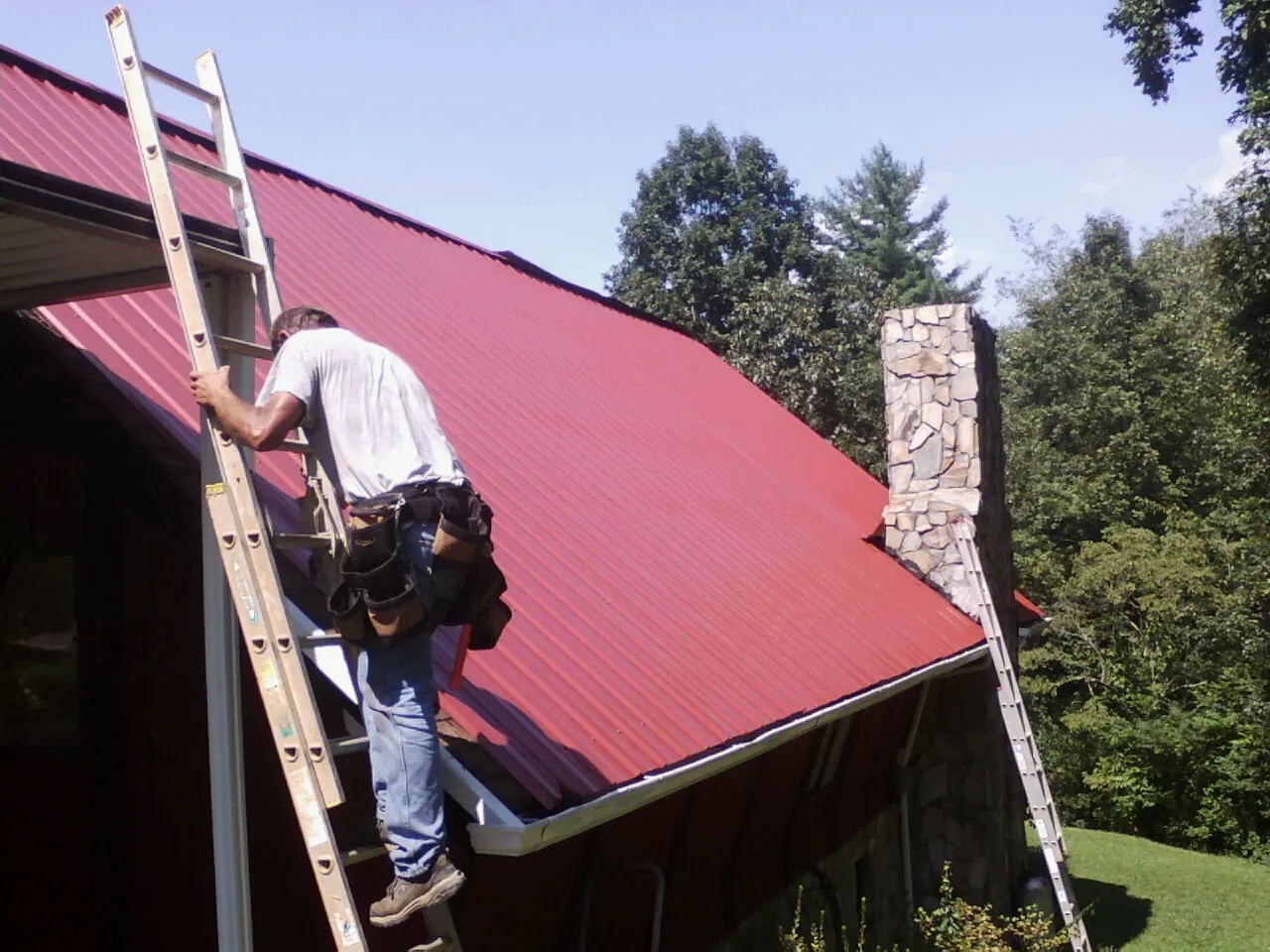 Roofer on a ladder beside a red metal roof and stone chimney — Soffit & Fascia Repair by Roofing Brothers in South Bloomingville